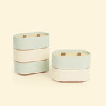 Bougies Totem S Vert d’eau – Citronnelle & Eucalyptus et Beige ivoire – Citronnelle & Citron, deux bougies ovales à double mèche en bois, posées sur un fond beige clair. 