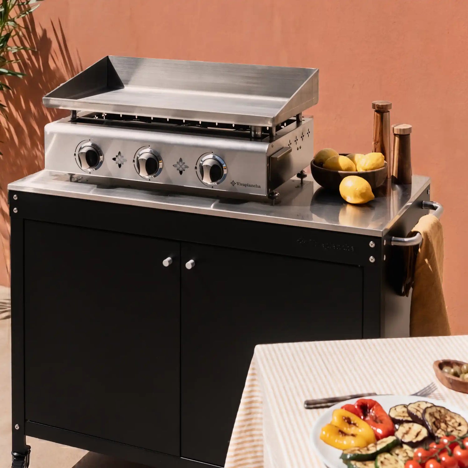 Plancha Vivaplancha sur la desserte Bodega avec assiette de légumes grillés au premier plan, en ambiance extérieure.
::visuel:avecportes