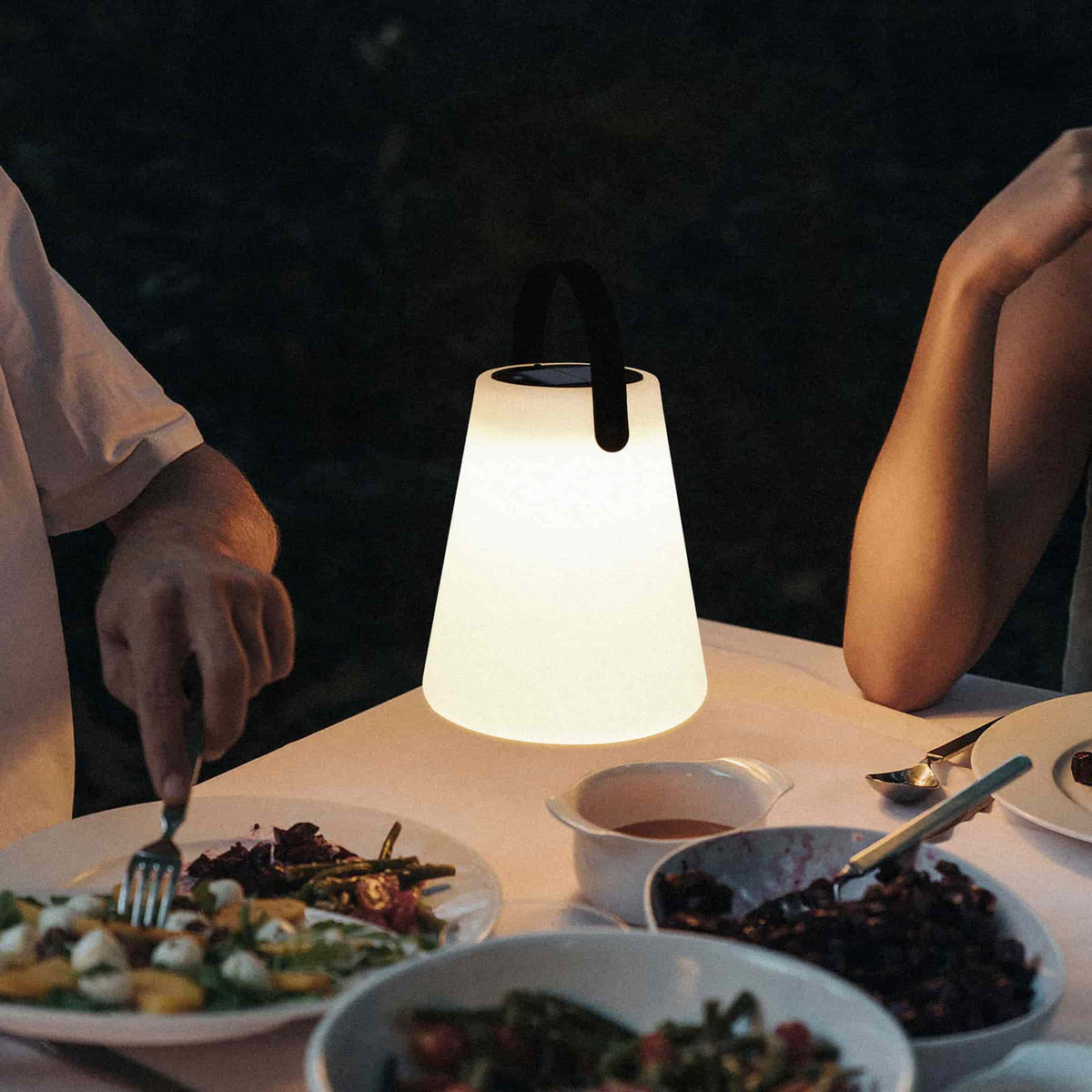 Lampe conique solaire allumée, diffusant une lumière chaude sur une table dressée pour un dîner en extérieur, entourée de plats et de convives.