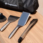 Vivaplancha utensils placed on a wooden worktop: wide spatula, long spatula, stainless steel tongs and cleaning ball, with the carrying bag visible in the background.