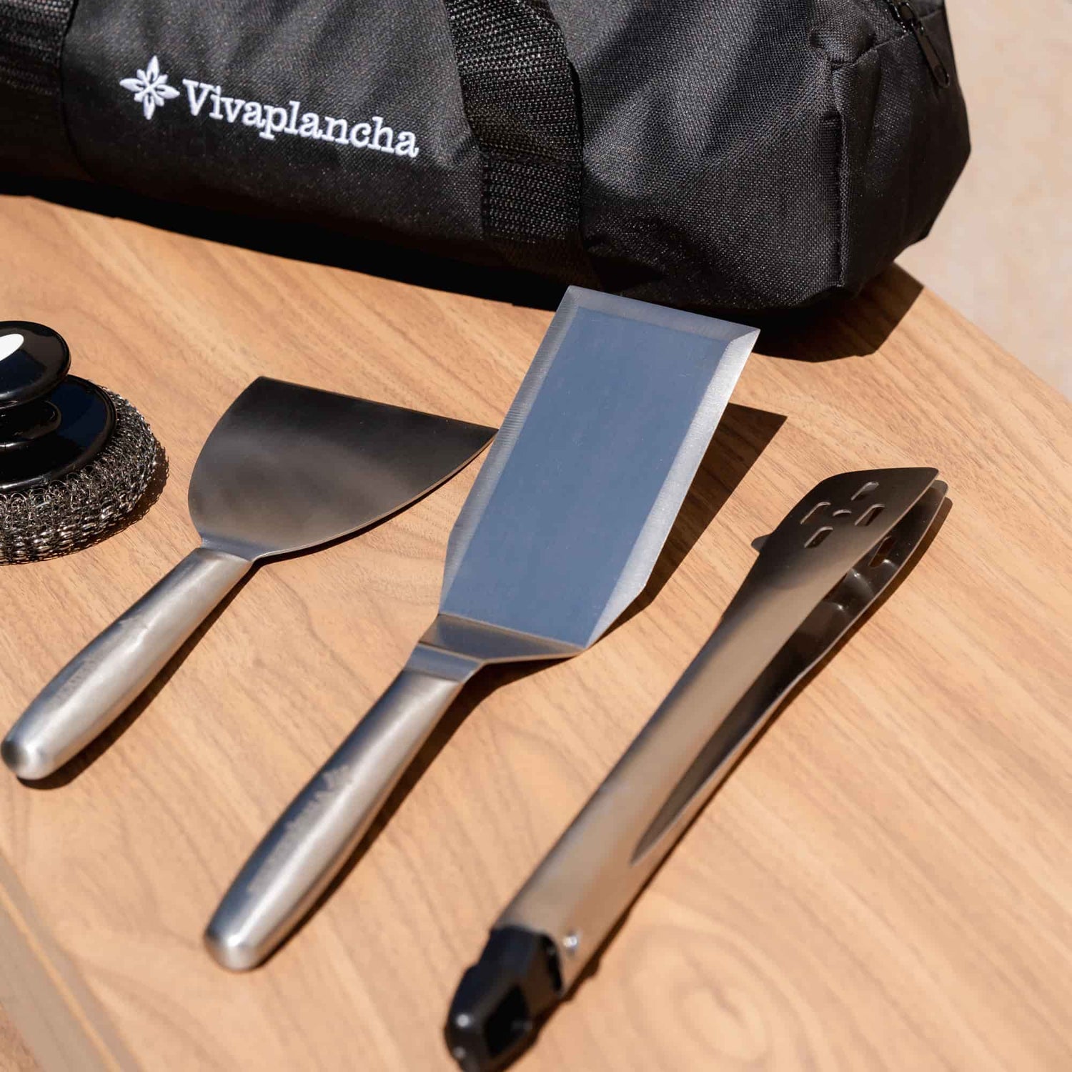 Vivaplancha utensils placed on a wooden worktop: wide spatula, long spatula, stainless steel tongs and cleaning ball, with the carrying bag visible in the background.