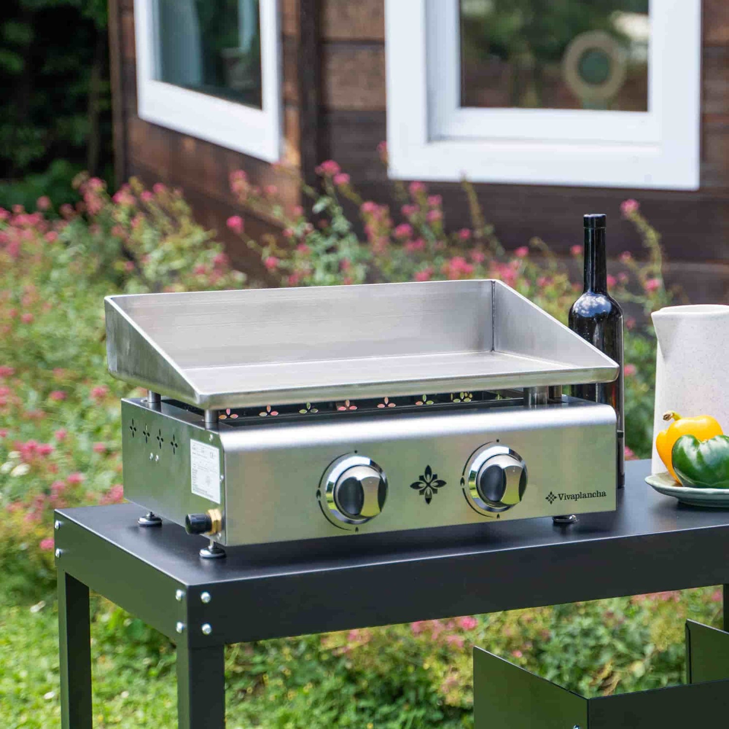 Plancha gaz Vivaplancha en inox posée sur une desserte extérieure noire, entourée d’un jardin fleuri.
