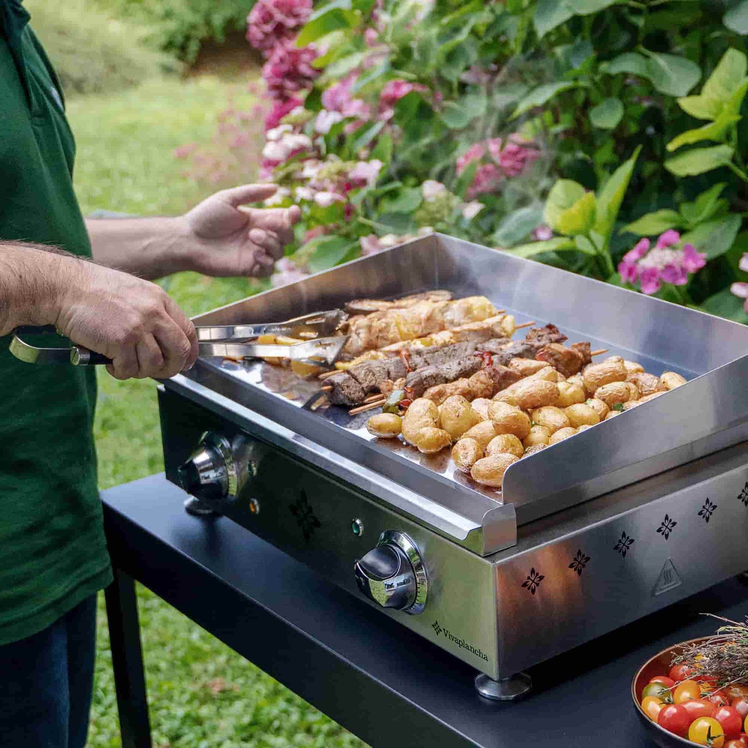 Personne cuisinant des brochettes et pommes de terre sur une plancha inox Vivaplancha en extérieur, avec de la vapeur s’élevant des aliments.