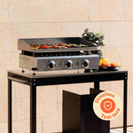 Vivaplancha stainless steel gas plancha in operation, with grilled vegetables on the plate and a black metal trolley, displayed outdoors with the “All stainless steel structure” icon.