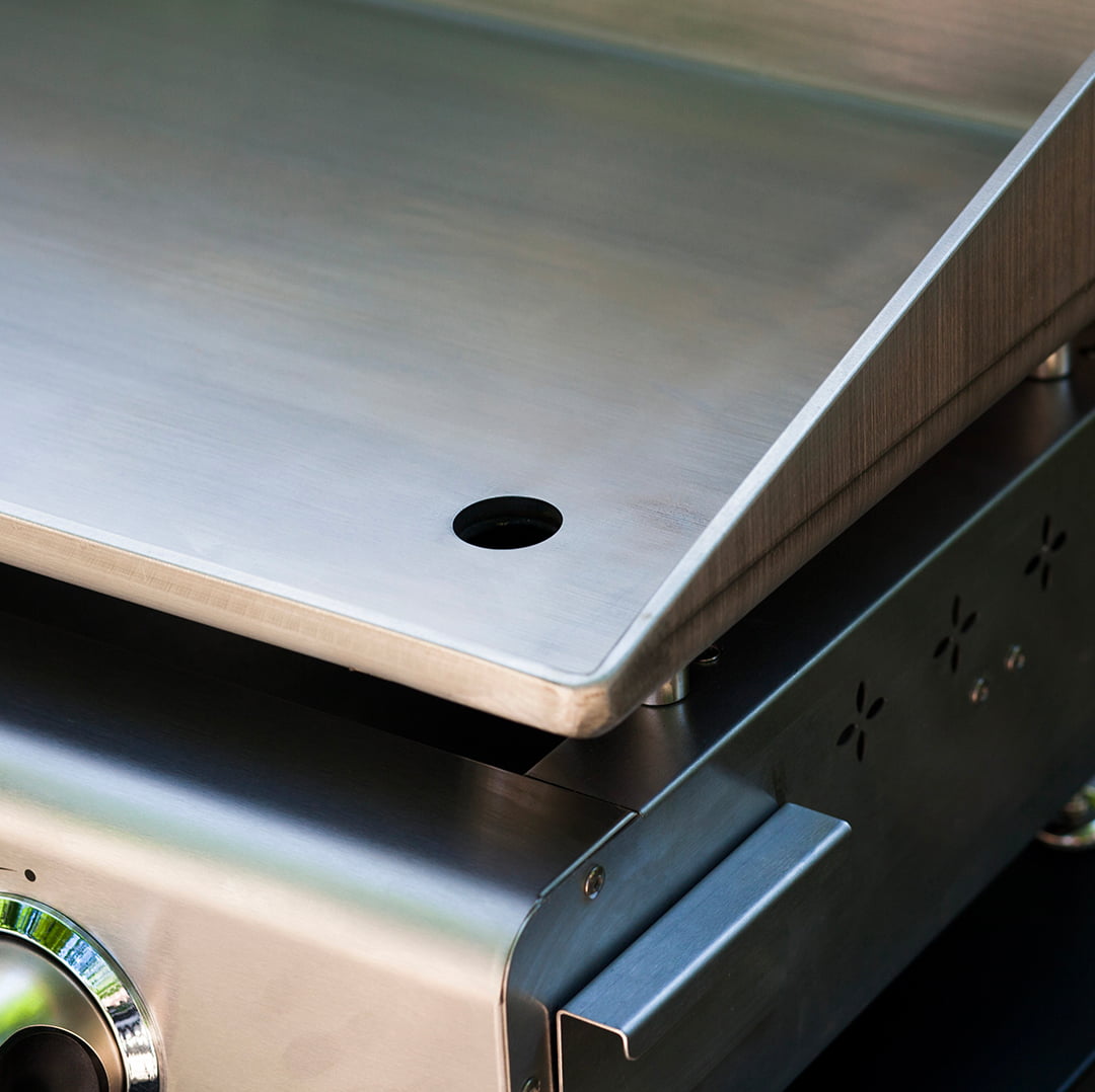 Gros plan sur la plaque de cuisson en inox d’une plancha Vivaplancha, montrant l’orifice d’évacuation des graisses et le bord relevé, avec la façade en inox visible en dessous.